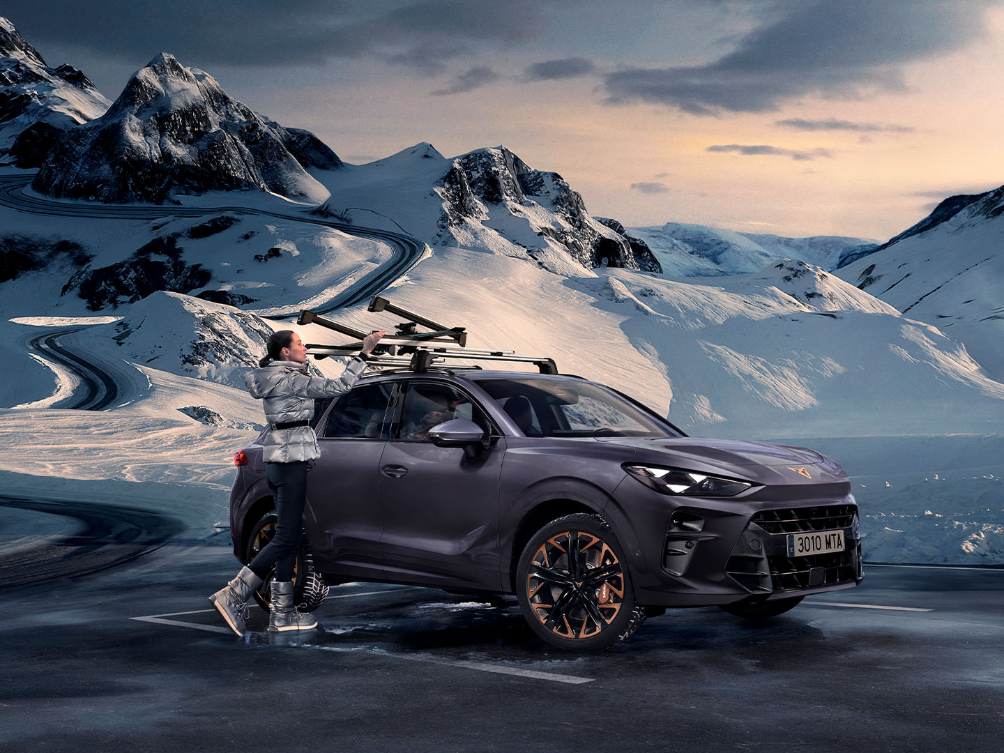 CUPRA Terramar lumisessa maisemassa, kuvassa yksi kuljetuksen lisävarusteista, suksiteline, joka on kiinnitetty auton katolle. Nainen käyttää telinettä suksien kiinnittämiseen.